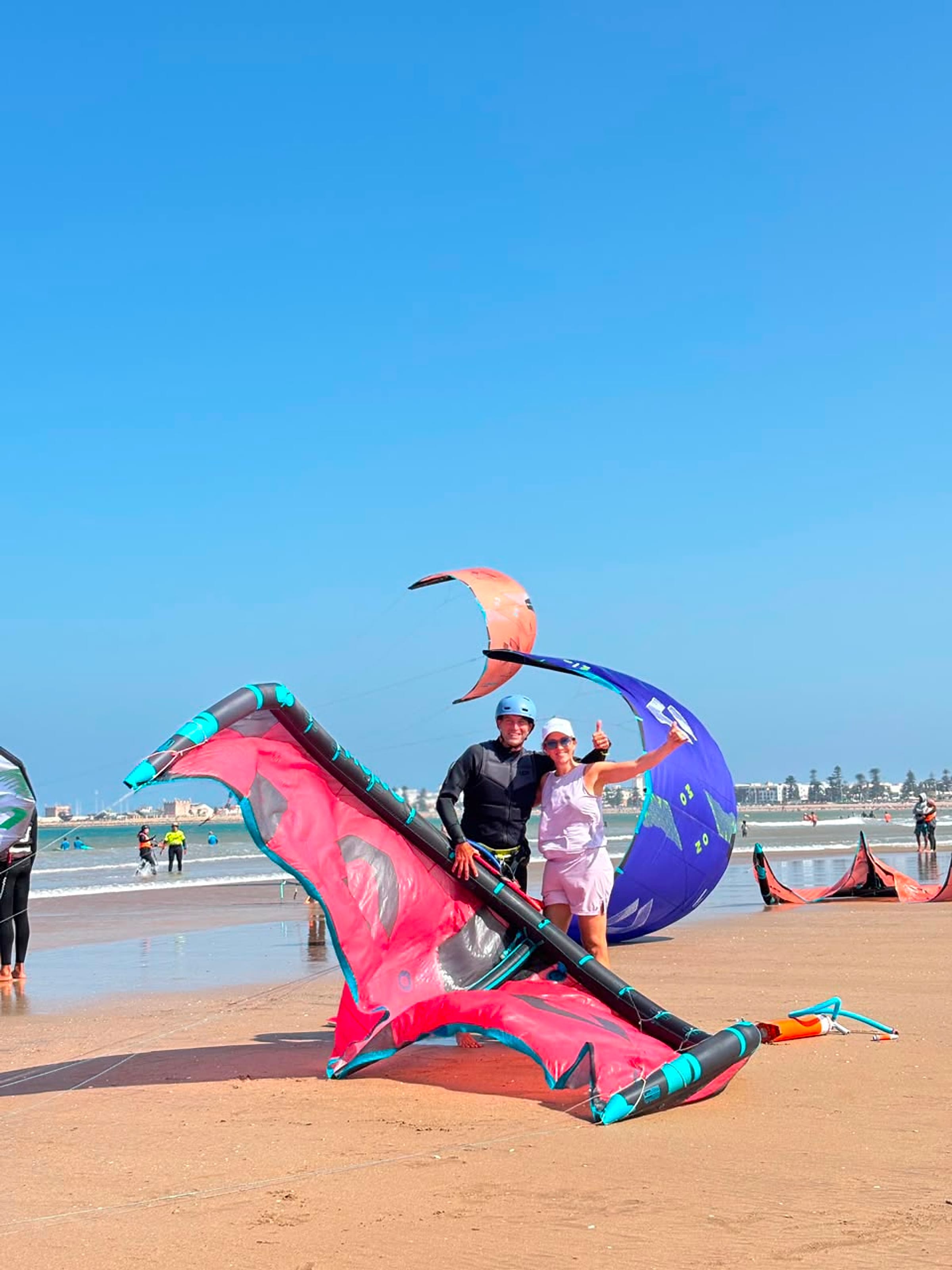 Two people stand among colourful kitesurf kites.